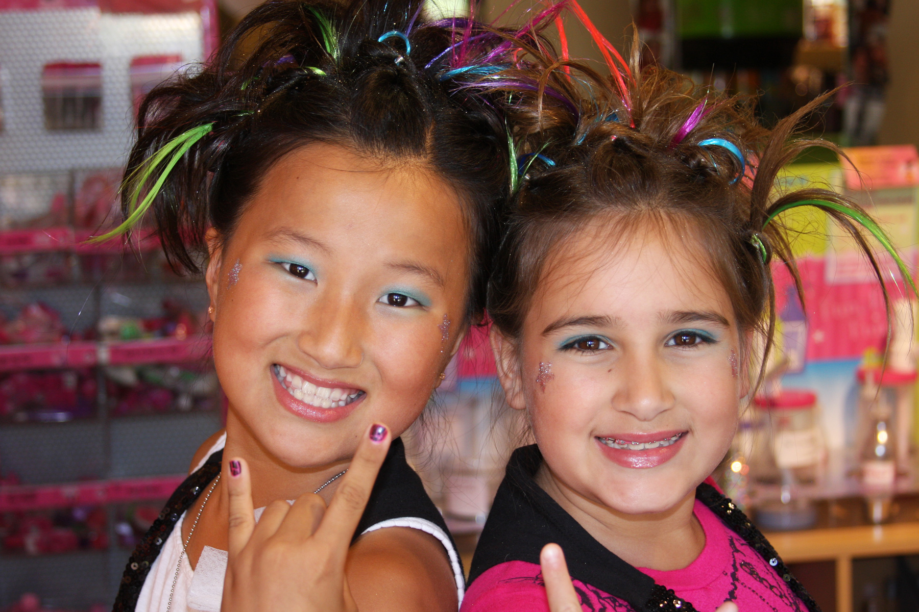 Two girls with face paint, makeup, and colored hair, smile and give hand gestures to the camera. | Sweet & Sassy